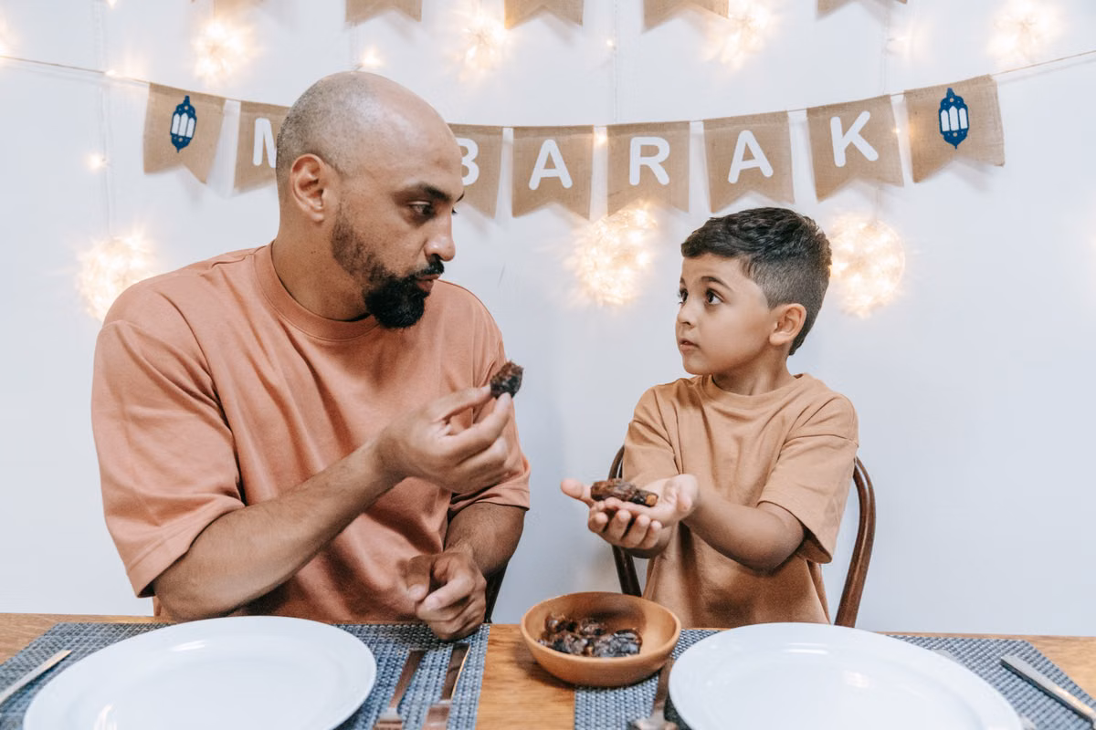 A father and son share dates during an Eid Mubarak celebration indoors.